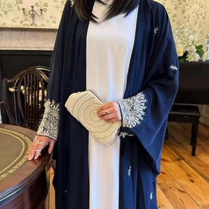 以下が含まれることがあります： 白のドレスの上に着用された、袖と縁に複雑な銀色の刺繍が施されたネイビーブルーのオープンアバヤ。真珠で覆われたクリーム色のクラッチバッグが手に持たれています。アバヤは繊細な刺繍が施されています。