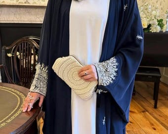 May include: A navy blue open abaya with intricate silver embroidery on the sleeves and edges, worn over a white dress. A cream-colored clutch covered in pearls is held in the hand. The abaya features delicate embroidered details.