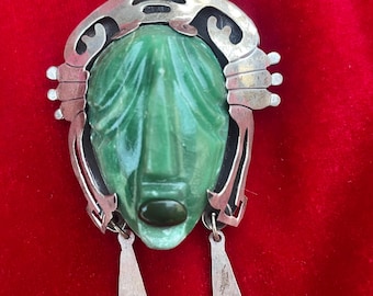 May include: A vintage brooch featuring a carved green face with a silver-tone metal frame. The face has detailed features and is accented with dangling silver-tone pendants. The brooch is set against a red background.