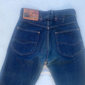 May include: Dark blue denim jeans featuring a high-waisted design with a tan leather patch on the back waistband. The jeans include two large back pockets with decorative stitching, a cinch back strap with metal hardware, and copper-colored rivets.