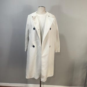 May include: A white double-breasted coat with black buttons. The coat is made of a textured fabric and has a long, flowing silhouette.