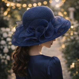 Op de afbeelding: Een marineblauwe zonnehoed met een brede rand en een groot, gerimpeld accent. De hoed is gemaakt van een geweven materiaal en heeft een decoratief, gelaagd stoffen detail. De vrouw op de foto draagt een bijpassende marineblauwe jurk.