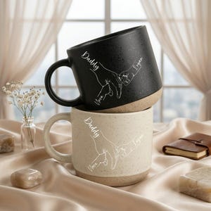 May include: Two ceramic mugs, one black and one cream, stacked on top of each other. The black mug has a speckled finish and a white graphic of two hands holding each other, with the words "Daddy", "Nova", and "Logan". The cream mug has a similar design.