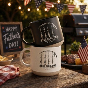 May include: Two ceramic mugs stacked on a wooden table. The top mug is black, the bottom is off-white. Both mugs have a fishing-themed design with the text "REEL COOL DAD" and names. A chalkboard with "HAPPY Father's DAY!" is in the background.