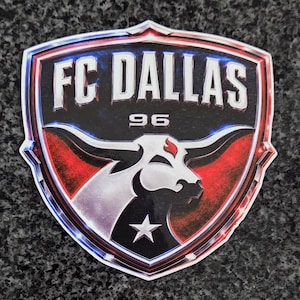 Puede incluir: Una pegatina en forma de escudo con el logotipo de FC DALLAS y el texto FC DALLAS y 96. El diseño incluye un gráfico de cabeza de toro blanca con un detalle de llama roja en el cuerno, una estrella blanca y un fondo negro con acentos rojos y azules.