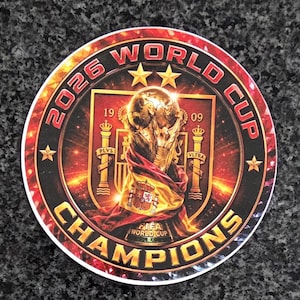 Op de afbeelding: Een ronde sticker met de FIFA Wereldbeker trofee gedrapeerd in een Spaanse vlag. Het ontwerp bevat de tekst 2026 WORLD CUP CHAMPIONS, twee gouden sterren en het Spaanse wapenschild met de jaartallen 1909 tegen een rode en zwarte achtergrond.