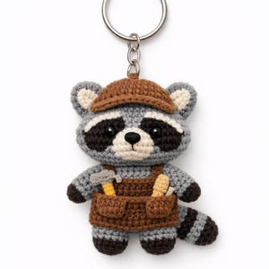 Puede incluir: Un llavero de mapache tejido a mano. El mapache es gris, negro y blanco, con un sombrero marrón y un peto. El peto tiene bolsillos con un martillo y una herramienta. El llavero tiene un anillo plateado.