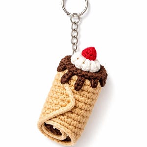 May include: A crocheted keychain shaped like a cannoli. The dessert-themed keychain features a tan-colored pastry with a dark brown chocolate drizzle, white cream, and a red crocheted strawberry on top. The keychain is attached to a silver metal ring.