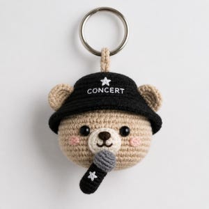 May include: A crocheted bear keychain with a black hat that says "CONCERT" and a small microphone. The bear is tan with pink cheeks, black eyes, and a brown nose. A silver key ring is attached to the top.