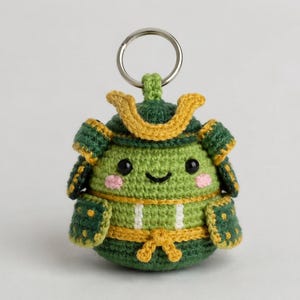 May include: A handmade crochet keychain shaped like a smiling samurai. The plush figure is mainly green with yellow and dark green details. It has a silver key ring and pink cheeks.