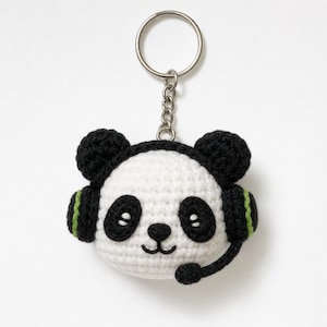 May include: A handmade crochet panda keychain with a headset. The panda has black and white coloring with black ears and eyes. The headset is black with green accents. The keychain is attached to a silver metal ring.