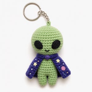 May include: A handmade green crochet alien keychain with black eyes and a smiling mouth. The alien wears a purple cape with yellow stars and pink dots. The keychain has a silver ring and chain.