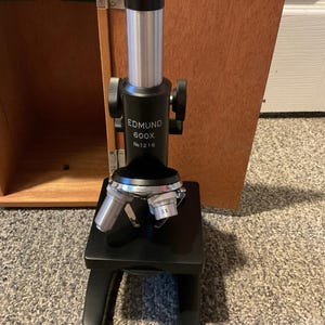 May include: A black Edmund 600X microscope with silver and black components. The microscope is set on a black base with curved legs. The wooden case is partially visible in the background. The microscope has the text "EDMUND 600X No 1216".