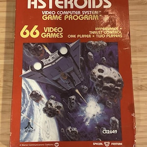 Puede incluir: Una caja de cartucho de videojuego Atari Asteroids vintage. La caja roja presenta el título del juego, un gráfico de nave espacial y el texto "66 Videojuegos". El texto adicional incluye "Hyperspace, Thrust Control, One Player, Two Players."