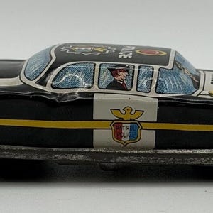 Könnte beinhalten: Ein altes Polizeiauto-Spielzeug aus Blech in Schwarz und Weiß mit einem gelben Seitenstreifen. Das Fahrzeug zeigt ein schildförmiges Emblem an der Tür mit der Aufschrift PATROL POLICE und eine grafische Darstellung eines Fahrers im Fenster.