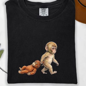 May include: Black t-shirt featuring a whimsical illustration of two monkeys. One monkey is walking, holding the hand of another monkey lying on the ground. The shirt has a Comfort Colors tag.