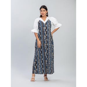 Puede incluir: Un vestido largo hasta el suelo con un corpiño blanco de cuello en V, mangas cortas abullonadas y una falda larga y recta. La falda muestra paneles verticales de tela azul y negra, cada uno decorado con patrones florales circulares y geométricos.