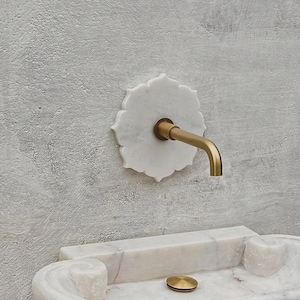 May include: A brass faucet with a white marble floral backplate, mounted on a textured grey wall. The spout curves downwards. A matching marble sink is visible below. The design is elegant.