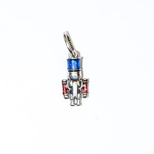 May include: A silver-toned charm in the shape of a toy soldier nutcracker. The nutcracker has a blue hat, red arms, and is detailed with the word "PANDORA". This charm is a festive accessory for bracelets or necklaces.