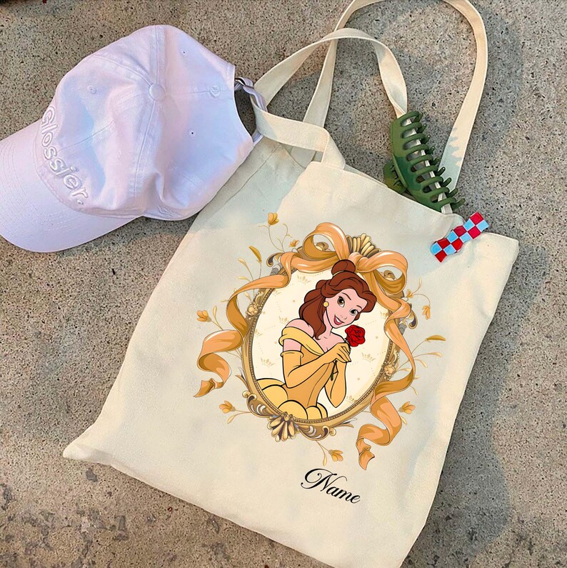 K&ouml;nnte beinhalten: Cremefarbene Tragetasche mit einer Illustration von Belle, die eine rote Rose h&auml;lt, goldfarben umrahmt, mit dem Wort "Name" darunter. Eine wei&szlig;e Baseballkappe und Haaraccessoires sind ebenfalls sichtbar.