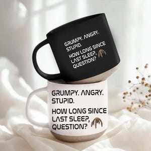 May include: A black mug stacked on a white mug, both featuring a speckled beige base. Each mug displays the text: GRUMPY. ANGRY. STUPID. HOW LONG SINCE LAST SLEEP, QUESTION? accompanied by a small brown graphic of a creature with multiple legs.