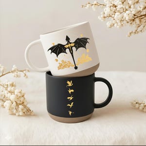 May include: Two ceramic mugs stacked. The top mug is white with a black dragon design and gold accents. The bottom mug is black with a series of gold dragon silhouettes. Both mugs have a tan base and a curved handle.