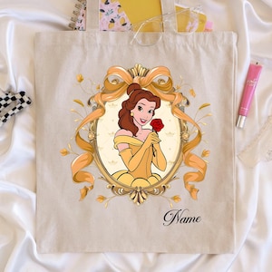 K&ouml;nnte beinhalten: Beige Tragetasche mit einer Cartoon-Illustration einer Frau in einem gelben Kleid, die eine rote Rose h&auml;lt, umrahmt von einem goldenen Band und floralem Design. Das Wort "Name" ist unten in Kursivschrift geschrieben.