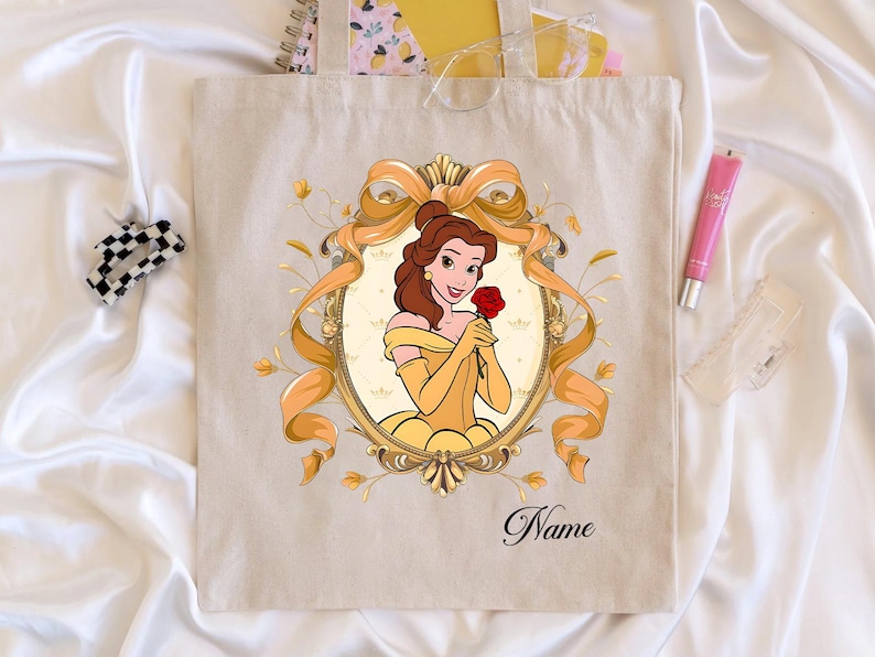 K&ouml;nnte beinhalten: Beige Tragetasche mit einer Cartoon-Illustration einer Frau in einem gelben Kleid, die eine rote Rose h&auml;lt, umrahmt von einem goldenen Band und floralem Design. Das Wort "Name" ist unten in Kursivschrift geschrieben.
