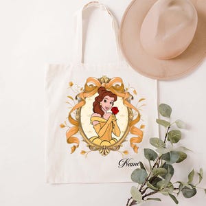K&ouml;nnte beinhalten: Cremefarbene Tragetasche mit einer Cartoon-Illustration einer Frau in einem gelben Kleid, die eine rote Rose h&auml;lt. Die Illustration ist in einem goldenen Oval mit einer goldenen Schleife und floralen Akzenten gerahmt. Das Wort "Name" ist auf der Tasche aufgedruckt.