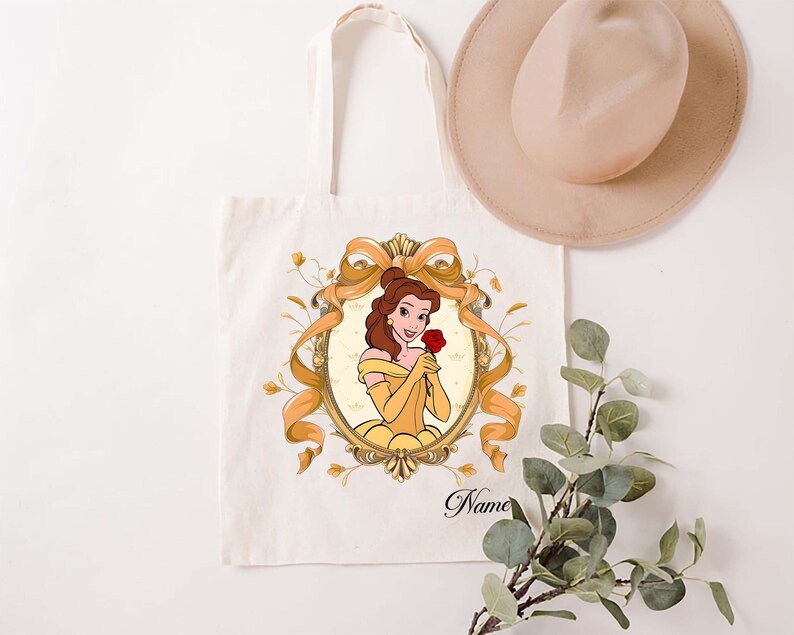 K&ouml;nnte beinhalten: Cremefarbene Tragetasche mit einer Cartoon-Illustration einer Frau in einem gelben Kleid, die eine rote Rose h&auml;lt. Die Illustration ist in einem goldenen Oval mit einer goldenen Schleife und floralen Akzenten gerahmt. Das Wort "Name" ist auf der Tasche aufgedruckt.