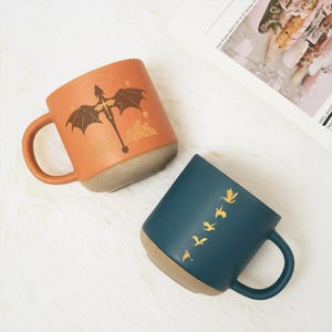 May include: Two ceramic mugs with a dragon theme. One is orange with a dragon illustration, the other is teal with a series of flying dragon silhouettes. Both mugs have a tan base and handle.
