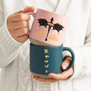 May include: A set of three ceramic mugs with a speckled, matte finish. The mugs are in shades of cream, light gray, and dark gray. Each mug has a simple, minimalist design and a rounded handle.