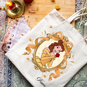 K&ouml;nnte beinhalten: Beige Tragetasche mit einer Cartoon-Illustration einer Frau in einem gelben Kleid, die eine rote Rose h&auml;lt. Die Illustration ist von einem goldfarbenen Oval und B&auml;ndern umrahmt. Das Wort "Name" steht unten in Schreibschrift.