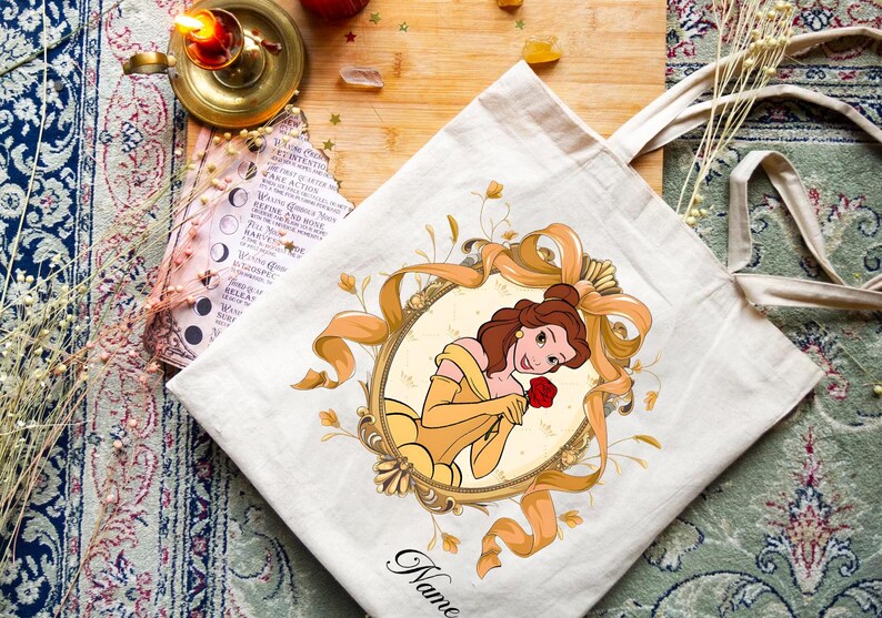 K&ouml;nnte beinhalten: Beige Tragetasche mit einer Cartoon-Illustration einer Frau in einem gelben Kleid, die eine rote Rose h&auml;lt. Die Illustration ist von einem goldfarbenen Oval und B&auml;ndern umrahmt. Das Wort "Name" steht unten in Schreibschrift.
