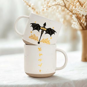 May include: Two white ceramic mugs stacked on top of each other. The top mug features a black dragon design with gold accents. The bottom mug has a series of gold bird silhouettes. Both mugs have a speckled finish and a tan base.