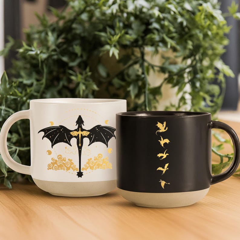May include: Two ceramic mugs with dragon designs. One mug is off-white with a black dragon and gold accents. The other is black with gold dragon silhouettes. Both mugs have a light beige base and a curved handle.