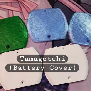 May include: A collection of Tamagotchi battery covers in various colors. The covers are in green, light blue, and white. The text "Tamagotchi (Battery Cover)" is printed on a black background. Each cover has a small hole for a screw.