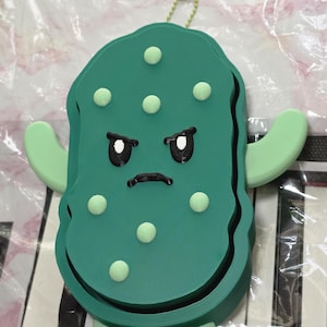 May include: A dark green, three-dimensional pickle figure with a grumpy facial expression, featuring light green raised bumps, two light green arms extending to the sides, and two light green feet at the base, topped with a small green ball chain.