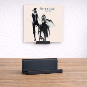 May include: A black vinyl record stand on a wooden surface, displaying a Fleetwood Mac "Rumours" album cover. The album cover shows a black and white image of two people. The stand is rectangular and has a small lip to hold the record.