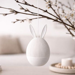 Op de afbeelding: Een wit, eivormig paasornament met verticale ribbels en konijnenoren. Het ornament hangt aan een dun touwtje, met een vage achtergrond met een vaas bloemen. Een decoratief item voor Pasen.