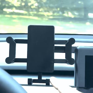 May include: Black car phone holder with adjustable arms and a cradle to secure a smartphone. The holder is mounted on a car dashboard, with a blurred view of the outside visible through the windshield.