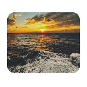 May include: Mousepad featuring a vibrant sunset over the ocean. The image shows a bright orange sun setting on the horizon, with golden clouds and dark blue water. White waves crash in the foreground, creating a dynamic seascape.