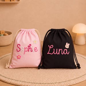 Op de afbeelding: Twee trekkoordtassen, één lichtroze en één zwart, elk voorzien van geborduurde namen in cursief schrift. De roze tas toont de naam Sophie met kleine bloemapplicaties, terwijl de zwarte tas de naam Luna toont met een kleine konijnenapplicatie.