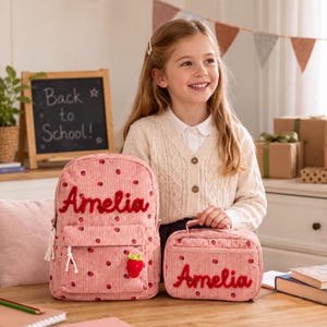 Könnte beinhalten: Ein rosafarbener Rucksack und eine passende Lunchtasche aus Kord, beide mit Erdbeermuster und dem in roter Schreibschrift gestickten Namen Amelia. Der Rucksack hat einen kleinen Erdbeeranhänger am Reißverschluss der Vordertasche.