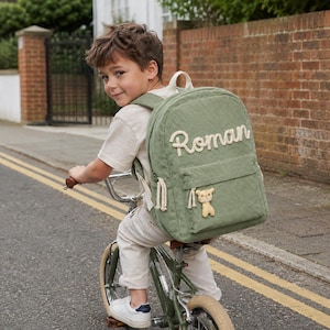 May include: A sage green corduroy backpack with the name "Roman" in white script. The backpack features a teddy bear applique and cream straps. The child is riding a bicycle.