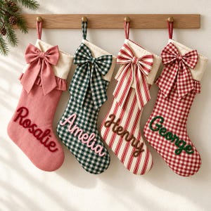 Op de afbeelding: Vier gepersonaliseerde kerstsokken hangend aan een houten rek. De eerste is effen roze met een strik en de naam Rosalie. De tweede is groen geruit met een strik en de naam Amelia. De derde is rood gestreept met een strik en de naam Henry. De vierde is rood geruit met een strik en de naam George.