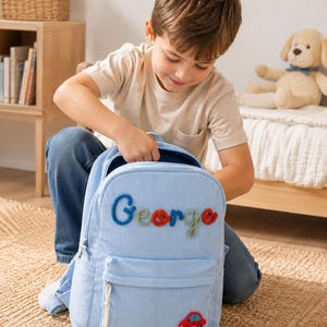 Könnte beinhalten: Hellblauer Cord-Rucksack mit dem Namen "George" in bunten Fäden gestickt. Auf der Vordertasche befindet sich ein kleiner roter Autoaufnäher. Der Rucksack hat eine Vordertasche und einen oberen Griff.