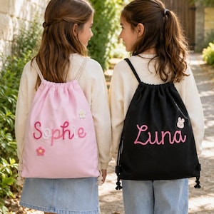 May include: Two drawstring backpacks, one light pink and one black, each featuring a personalised name embroidered in pink script. The pink bag displays the name Sophie with small flower accents, while the black bag features the name Luna with a small bunny icon.