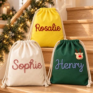 May include: Three drawstring bags in yellow, cream, and dark green. The yellow bag features the name Rosalie in red embroidery. The cream bag displays the name Sophie in red and white striped embroidery. The green bag shows the name Henry in blue and white striped embroidery with a small reindeer applique.
