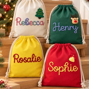Könnte beinhalten: Vier Geschenktaschen mit Kordelzug aus cremefarbenem, grünem, gelbem und rotem Stoff. Jede Tasche ist mit einem personalisierten Namen in strukturierter Schrift und einer kleinen weihnachtlichen Applikation wie Tannenbaum, Rentier oder Glocke versehen.
